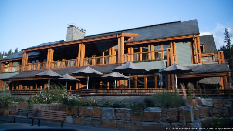 Timber-trimmed lodge-style building with balconies, umbrellas, stone landscaping, and a street-level terrace.