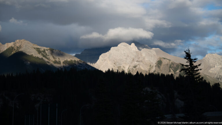 Sunlit pale limestone peaks rising above a dark conifer silhouette beneath heavy gray and white clouds near Banff.