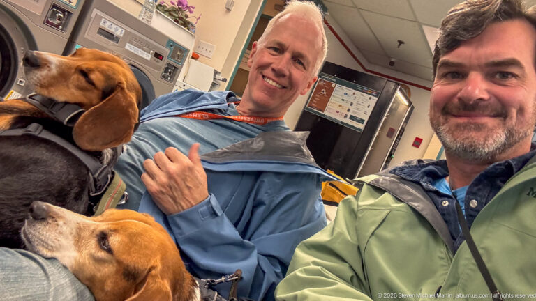 Two seated men smiling beside two beagles in harnesses inside a compact public waiting area.