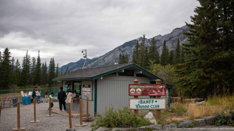 Banff Canoe Club rental kiosk with visitors at the service window, canoe signage, conifers, and mountains behind.