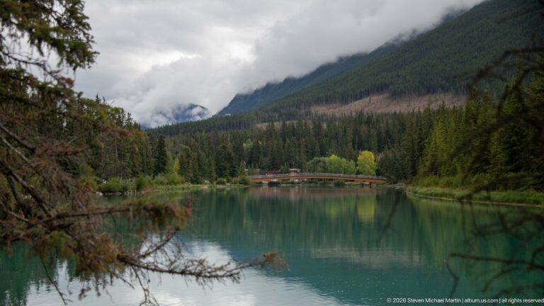 Calm green-blue water reflecting forested mountains, with a low bridge in the distance beneath cloud-covered slopes.