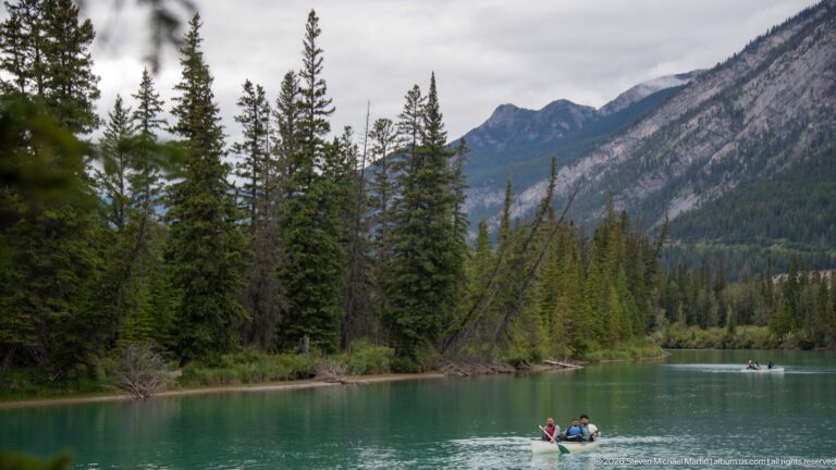 Two paddlers in a canoe on calm turquoise water, with conifer forest, mountains, and another distant craft.