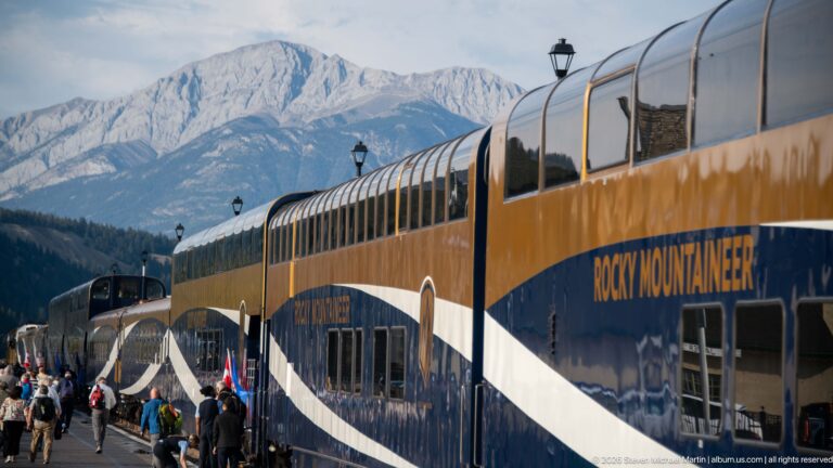Rocky Mountaineer glass-dome cars line a busy Banff platform with passengers and steep mountains rising behind them.