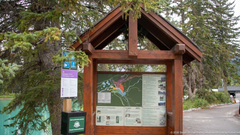 Timber information kiosk with large Banff-area map and visitor panels beside turquoise water and forest.