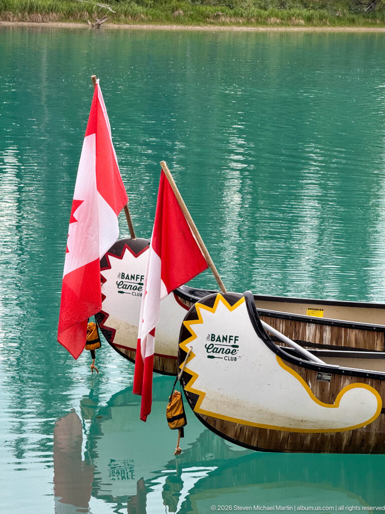 Close view of decorated Banff Canoe Club canoe bows and Canadian flags reflected in turquoise water.
