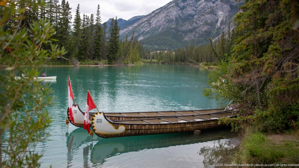 Decoratively painted Banff Canoe Club canoes with Canadian flags moored on turquoise water below forested mountains.