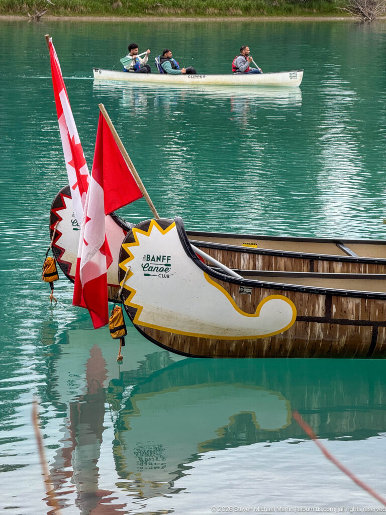 Two decorated Banff Canoe Club canoes with Canadian flags at a dock, with paddlers crossing turquoise water behind.