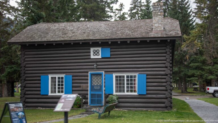 Dark log cabin with stone chimney, white multi-pane windows, bright blue shutters and door, and interpretive panels.