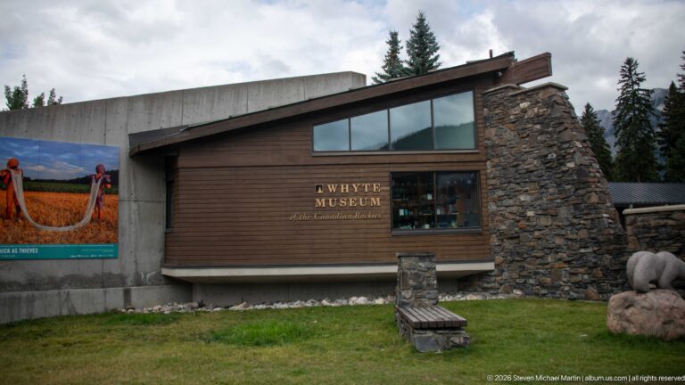 Whyte Museum façade with dark wood, concrete, broad windows, stone chimney, lawn, bench, and animal sculpture.