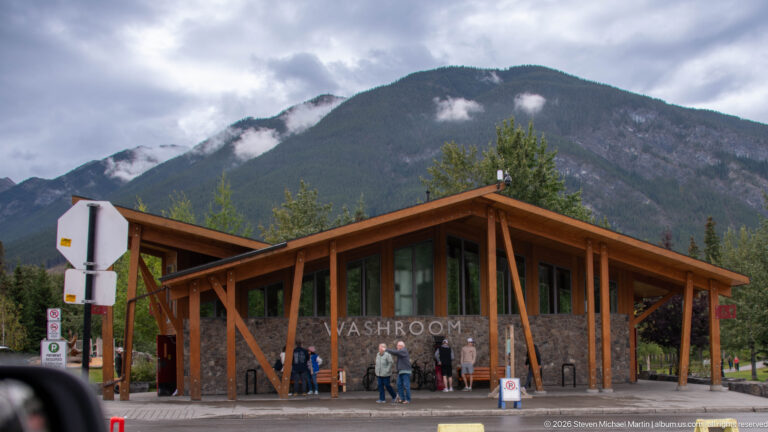 Modern Banff washroom with timber posts, stone walls, glass entrance, visitors, and a cloud-wrapped mountain behind.