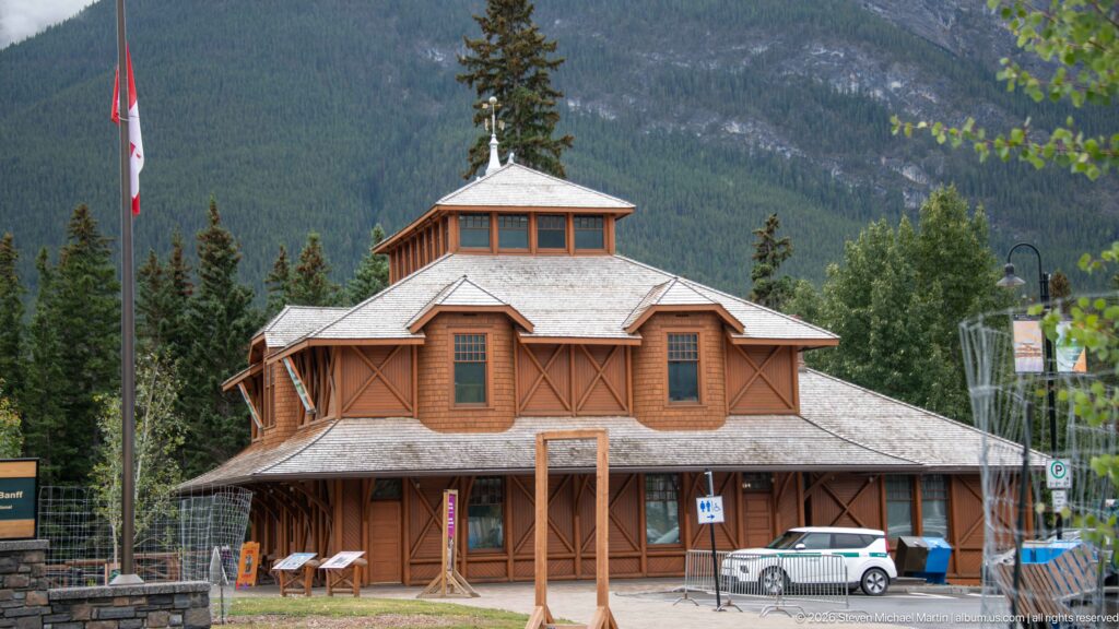 Side or rear elevation of Banff Park Museum with hipped roof, raised monitor, timber panels, fencing, and mountain slope.