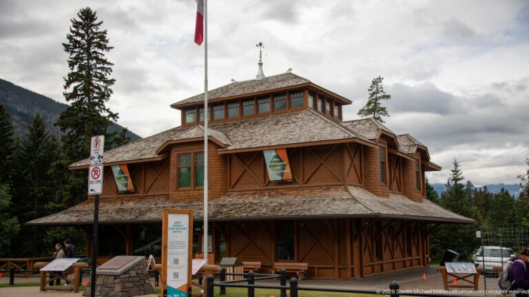 Corner of Banff Park Museum showing timber bracing, wraparound veranda, layered shingle roofs, flagpole, and interpretive signs.