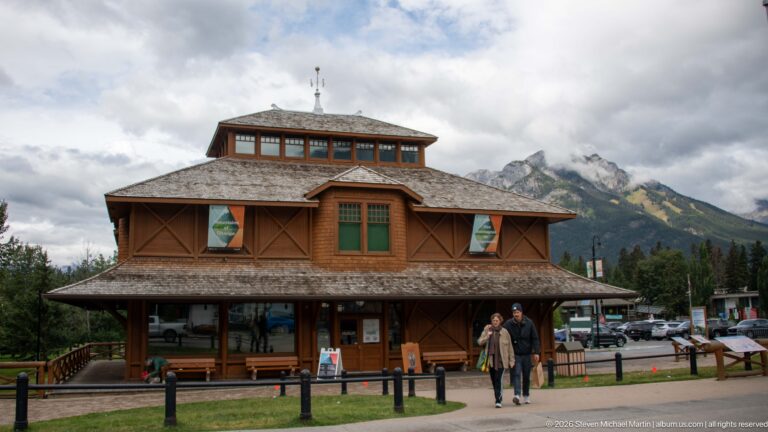 Brown timber Banff Park Museum with broad veranda, shingled roof, central clerestory, visitors, and mountains behind.