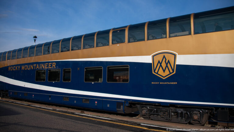 Side view of a navy, white, and gold Rocky Mountaineer glass-dome passenger railcar beside the platform.
