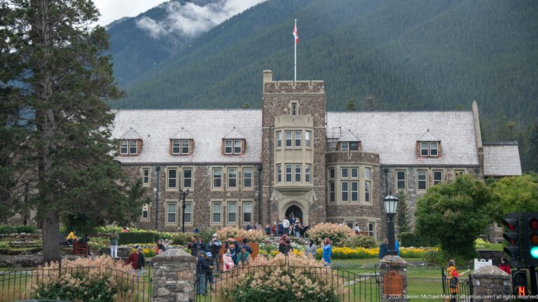 Front façade of Banff Park Administration Building beyond flower gardens, visitors, and a Canadian flag.