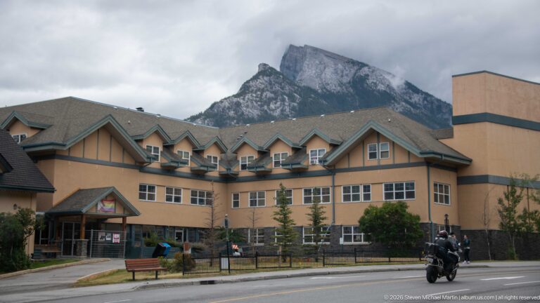 Tan multi-story Banff building with gabled dormers and balconies below a steep mountain ridge under gray clouds.