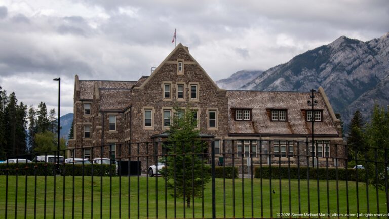 Side view of the stone Banff Park Administration Building across lawn and iron fencing, with mountains behind.