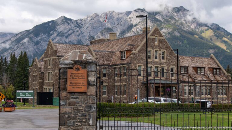 Stone entrance pier and iron fence before the gabled Banff Park Administration Building, with cloudy mountains behind.