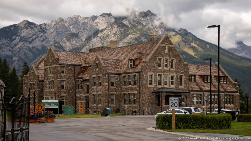 Large stone Banff Park Administration Building with steep gables and multi-pane windows beneath a rugged mountain ridge.