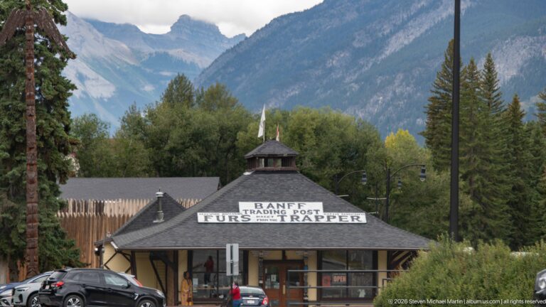 Rustic Banff Trading Post with “Furs & Trapper” roof sign, parked cars, trees, and mountains behind.