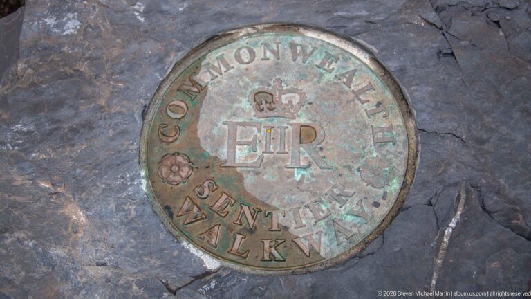 Weathered circular Commonwealth Walkway medallion with crowned ER monogram embedded in dark stone paving.