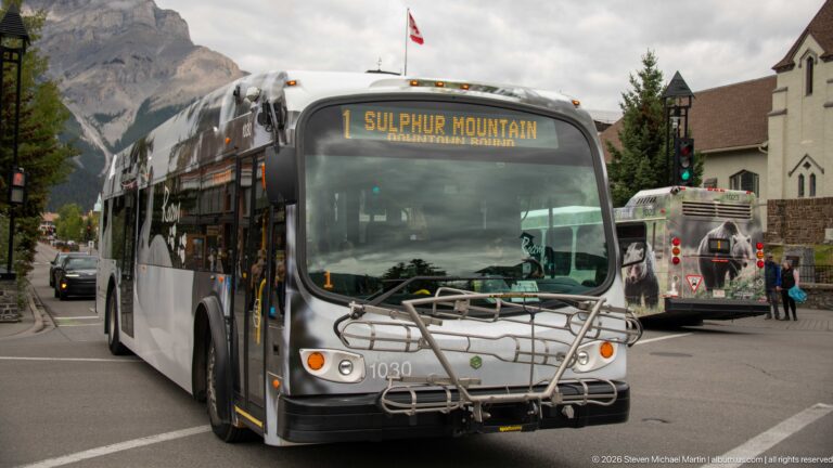 Roam transit bus marked Route 1 Sulphur Mountain on a Banff street, with storefronts and another bus behind.