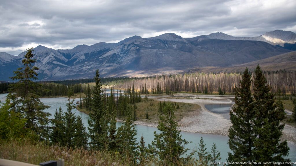 Turquoise braided river curves around gravel bars and conifer islands beneath a broad layered mountain ridge.