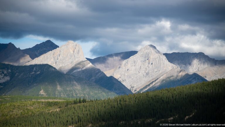 Two pale limestone peaks glow in sunlight above dark forest beneath heavy gray clouds.