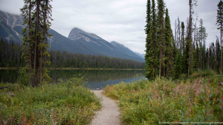 Gravel path curves through shrubs and conifers beside a mountain lake with steep peaks beyond.