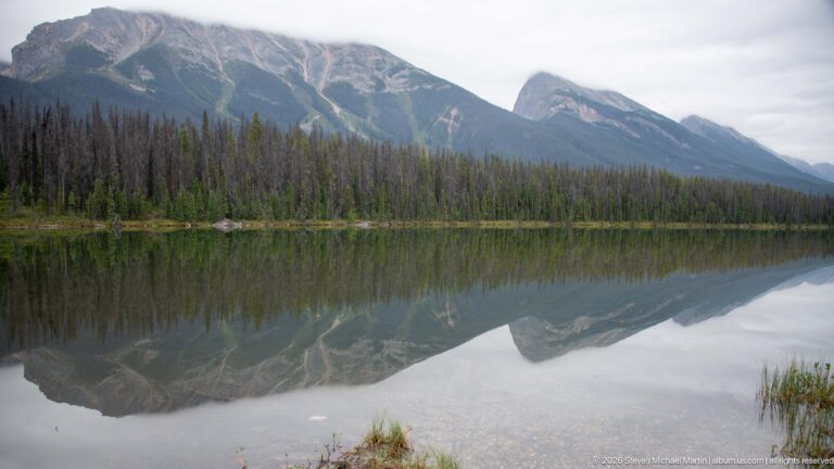 Rugged mountain ridge and forested slopes form a broad mirror reflection across a calm lake.