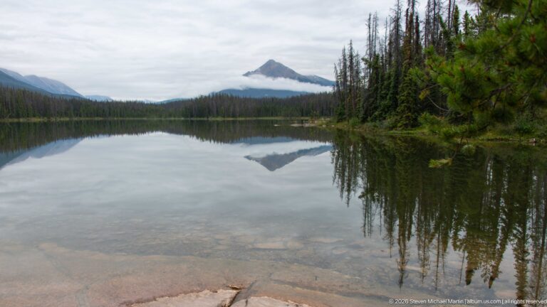 Triangular mountain and cloud band reflected symmetrically in a calm forest lake with clear shallows.