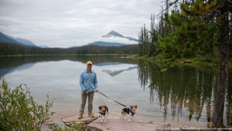 Man and two beagles stand on lakeshore rock with a triangular mountain reflected in calm water.