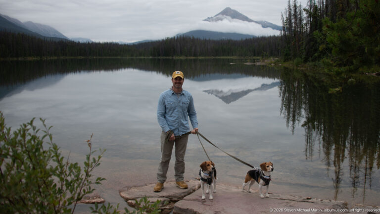 Man with two leashed beagles stands on a rocky shore before a reflective mountain lake.