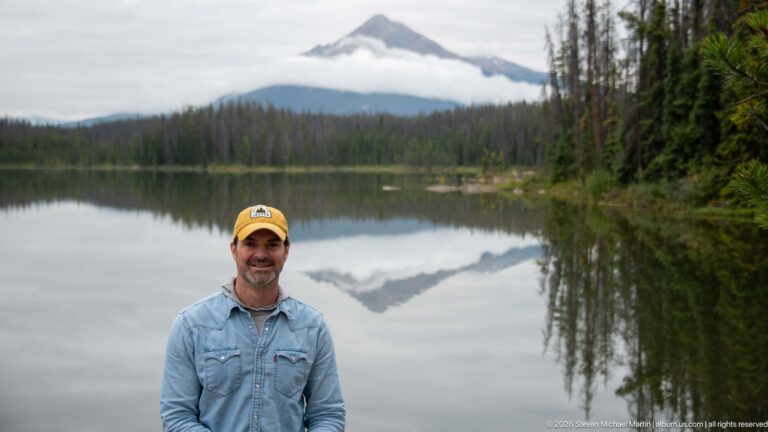 Man in a yellow cap stands before a calm lake reflecting a triangular mountain and low cloud band.