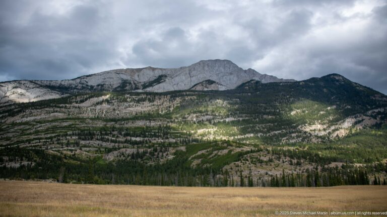Pale limestone ridge rises above sparsely forested slopes as sunlight breaks through dark storm clouds.