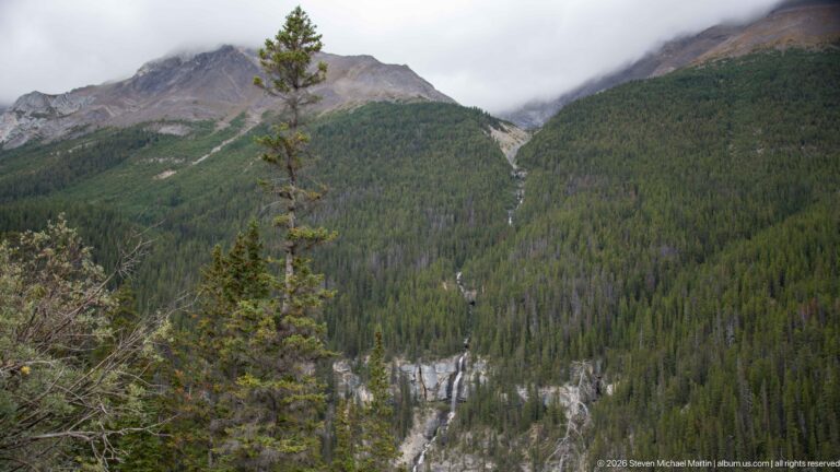 Thin waterfall drainage descends a broad forested mountainside beneath low clouds.