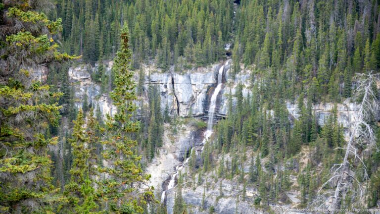 Long multi-stage waterfall descends through pale limestone cliffs and dense conifer forest.