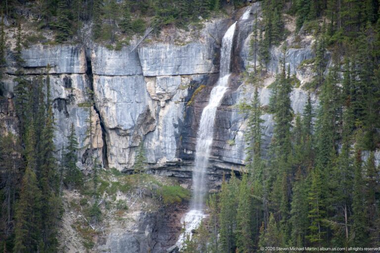 Tall narrow waterfall drops over a layered limestone cliff framed by evergreen trees.