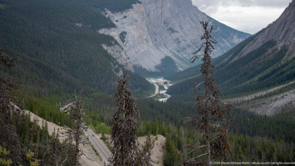 Highway descends through conifer forest into a steep mountain valley with a winding river below.