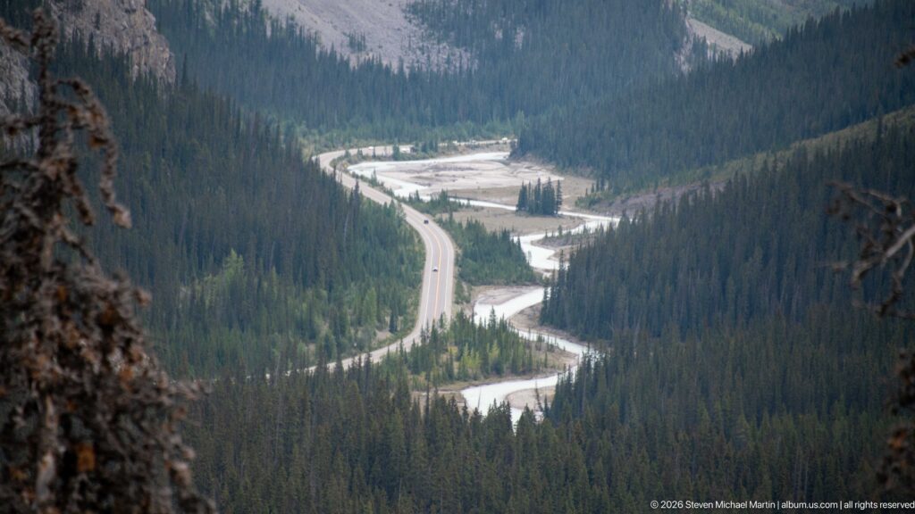 Curving highway parallels a pale braided river through a densely forested mountain valley.