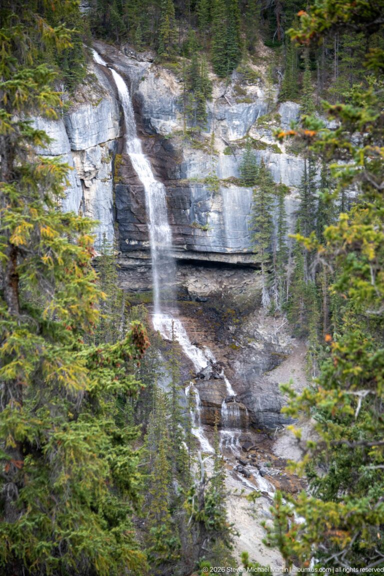 Multi-tiered waterfall descends a gray limestone cliff surrounded by dense evergreen forest.