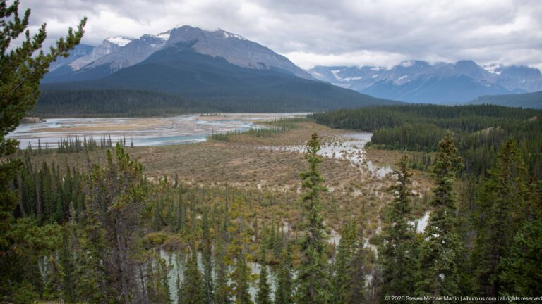 Braided river and gravel floodplain extend through conifer forest toward cloud-covered mountain ranges.