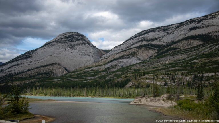 Turquoise river bends around a gravel point below steep folded limestone mountains and dark conifer bands.