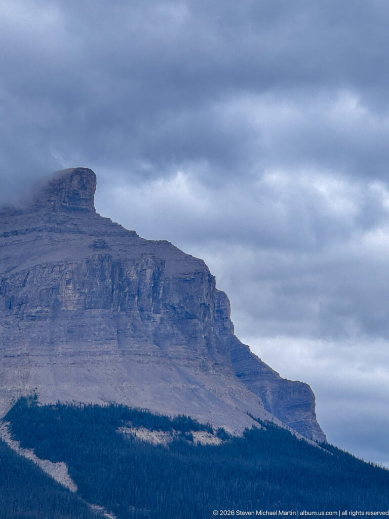Flat-topped layered limestone mountain rises above conifer forest beneath dark storm clouds.