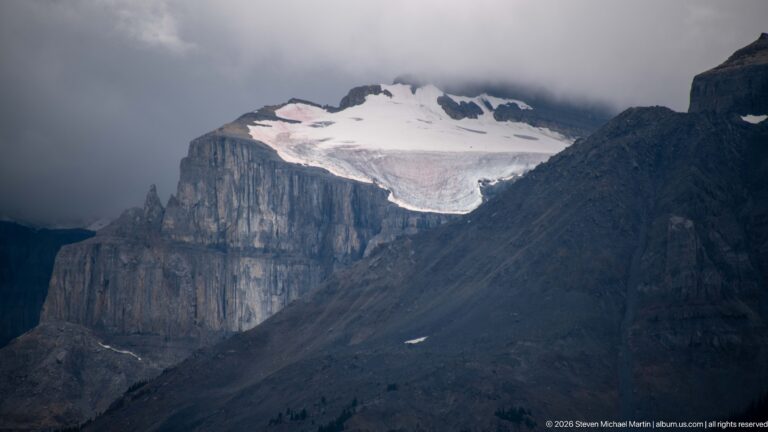 White snow or ice field lies above sheer layered limestone cliffs beneath dark low cloud.