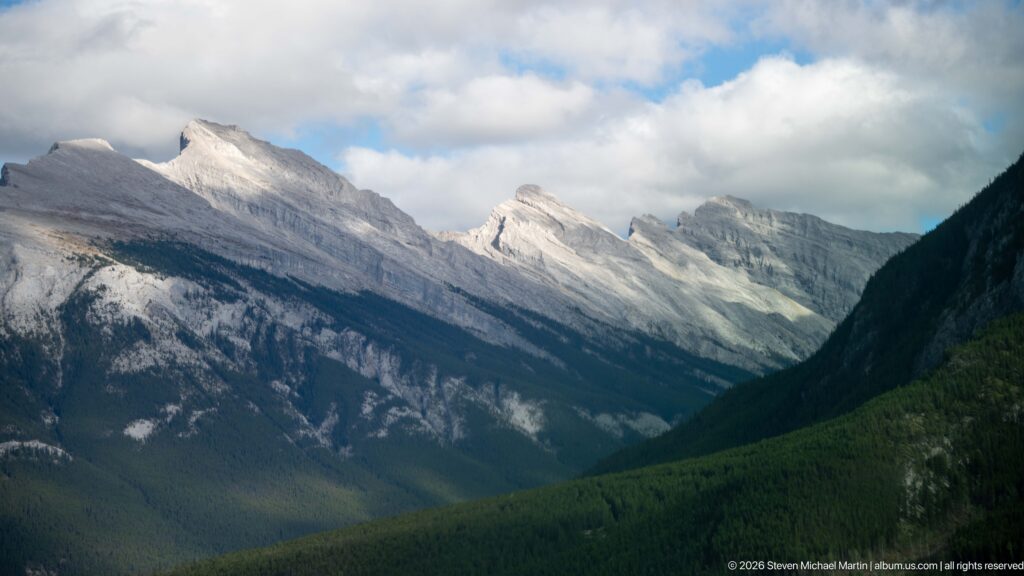 Several pale, tilted limestone peaks recede above a dark green forested valley beneath broken clouds.