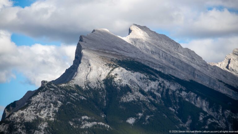 Steep pale limestone ridge with tilted rock layers rises above dark forest under broken clouds.