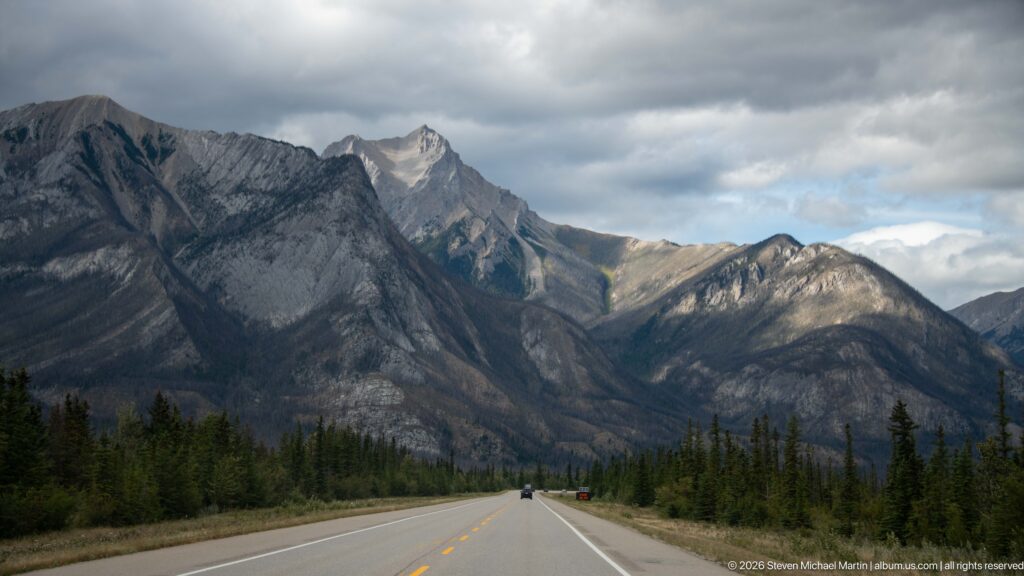 Straight mountain highway leads between conifer forest toward steep gray peaks beneath broken storm clouds.