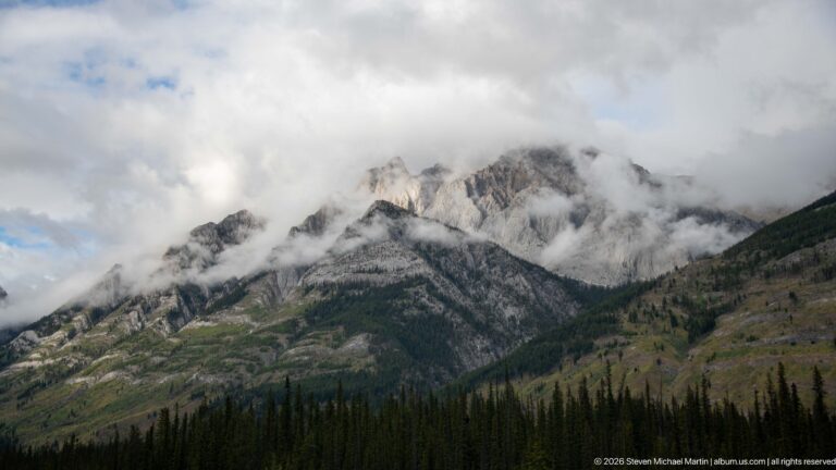 Jagged pale limestone ridge rises above dark conifers as clouds drift through its gullies and summits.