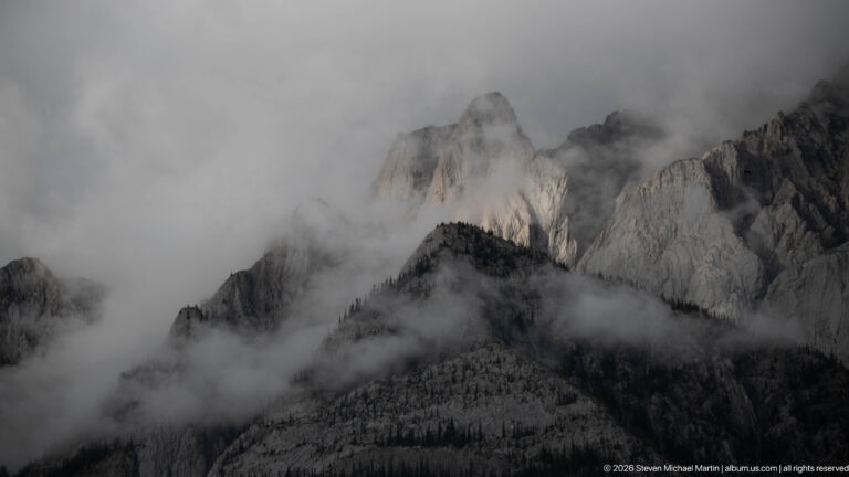 Pale jagged limestone towers emerge through dense gray cloud above a dark forested slope.